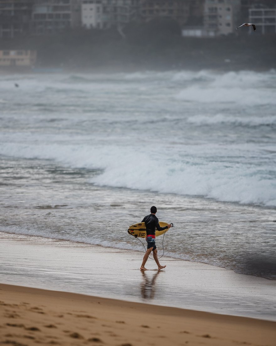 A surfer carries a yellow surfboard beside rolling waves.