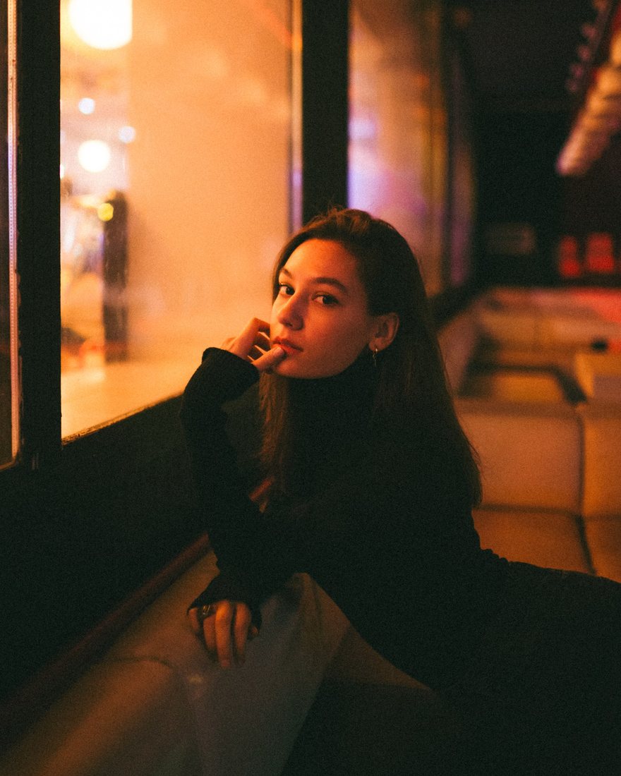 A woman rests beside a warmly lit window at night.