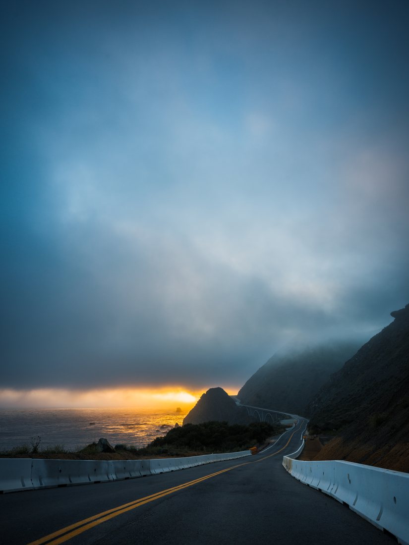 A coastal road curves beneath misty cliffs as golden sunlight breaks through the clouds over the ocean.