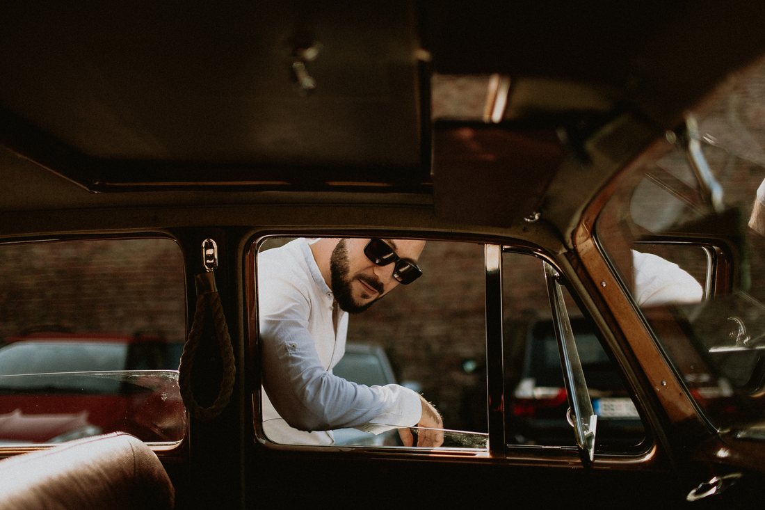 A portrait of a man wearing sunglasses, framed through the window of a vintage car.