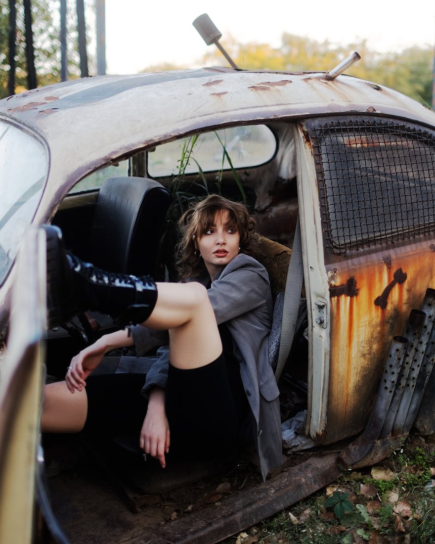 A woman in a grey jacket sits in the doorway of a weathered, rusted car.